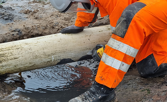 A man wearing high vis clothing and breathing apparatus using a hand power tool to clean a pipe in the sand