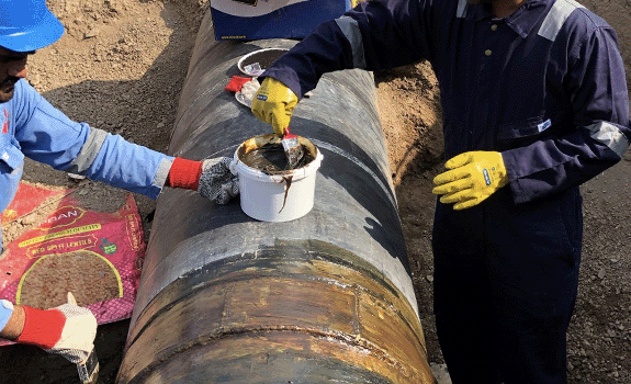 Brown paste being applied to a buried pipeline by workers in personal protective equipment.