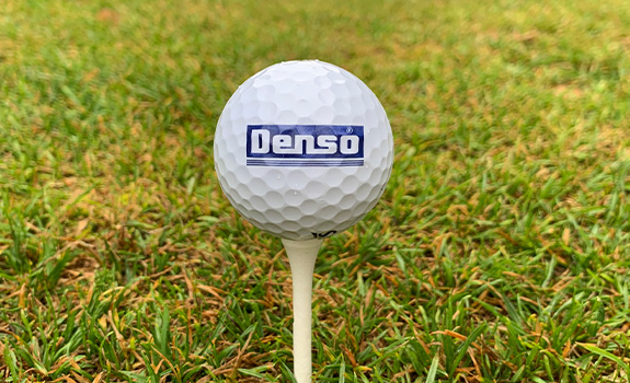 A Denso logo on a white golf ball, balancing on a white tee on some grass.
