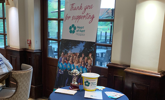 A charity pull up banner, collection bucket, and silver golf trophy on a blue table.