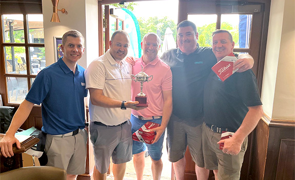 Five gentlemen posing for a picture with a golf trophy and other prizes.