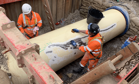 A pipe in a trench being washed by a worker in high vis and PPE.