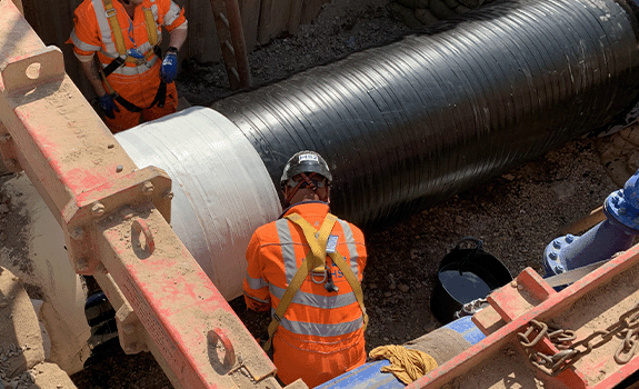 A pipe in a trench being wrapped by two workers in high vis and PPE.