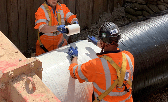 A pipe in a trench being wrapped by two workers in high vis and PPE.