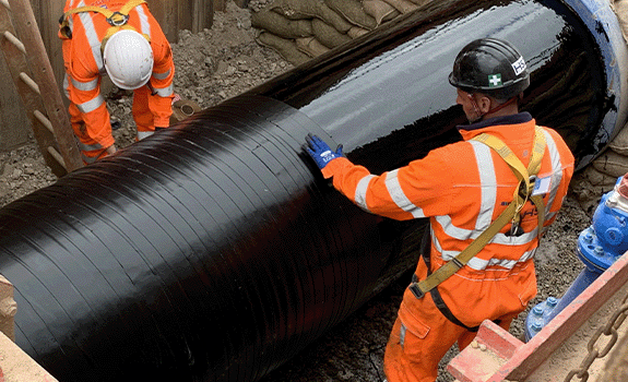 A pipe in a trench being wrapped with a black tape (Densoclad 70) by two workers in high vis and PPE.