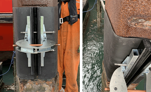 A black plastic sleeve (SeaShield 2000FD HDPE Jacket) being closed around a jetty pile with the help of a large metal pincer (SeaShield Clamping Tool)