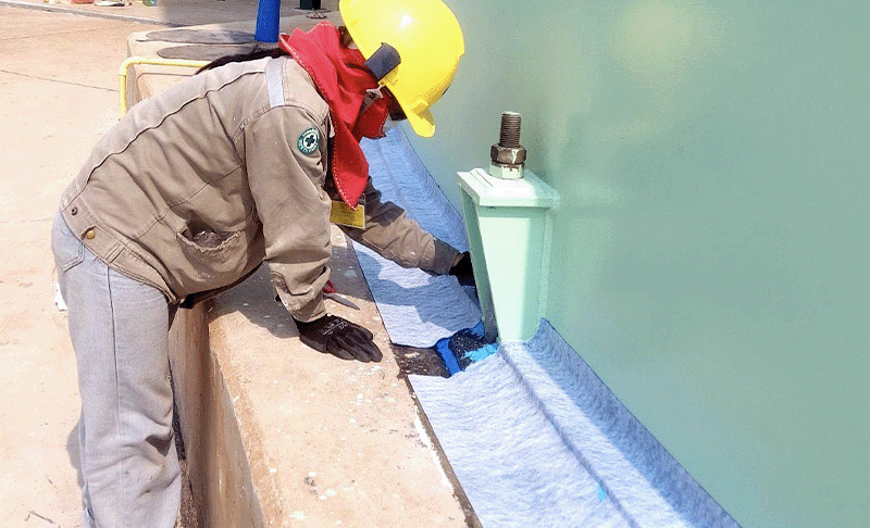 A worker in full PPE applying Viscotaq EZ Wrap (a blue fabric-backed tape) around an anchor bolt of a green aboveground storage tank sitting on a concrete foundation slab.