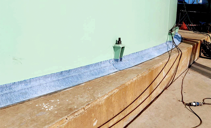 Viscotaq EZ Wrap (a blue fabric-backed tape) has been applied around the base of a green storage tank that sits on a sandy-coloured concrete foundation slab.