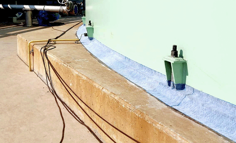 Viscotaq EZ Wrap (a blue fabric-backed tape) has been applied around the base of a green storage tank that sits on a sandy-coloured concrete foundation slab.