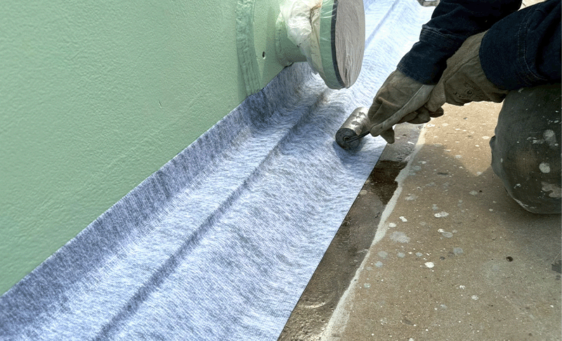 A worker in PPE gloves uses a roller to smooth out a blue fabric backed tape that has been applied between a green storage tank wall and the concrete floor.