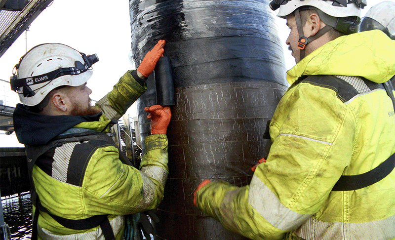 Two workers in high vis clothing and full PPE applying a black tape (Denso Glass Outerwrap) around a jetty pile, on top of a brown tape (Denso Marine Piling Tape).