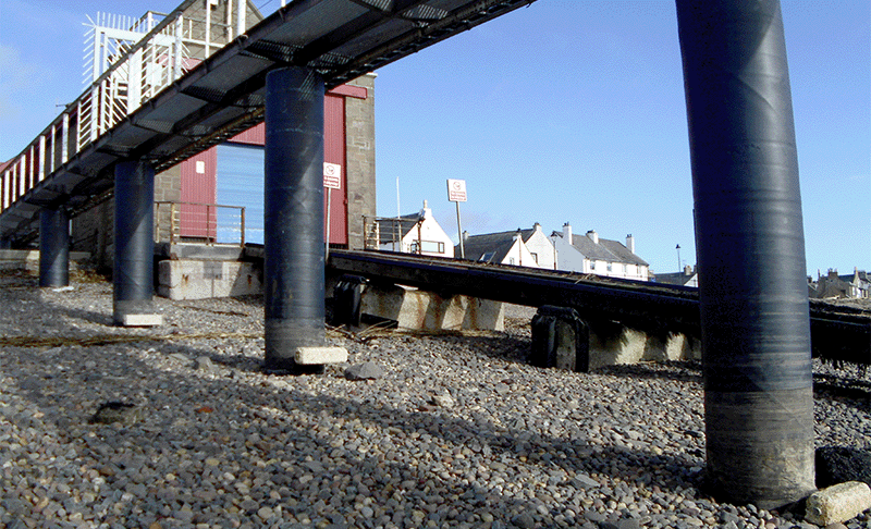 A raised walkway over a stony beach with support piles wrapped in a black protective jacket, set against a building with red doors and a ramp leading across the beach.